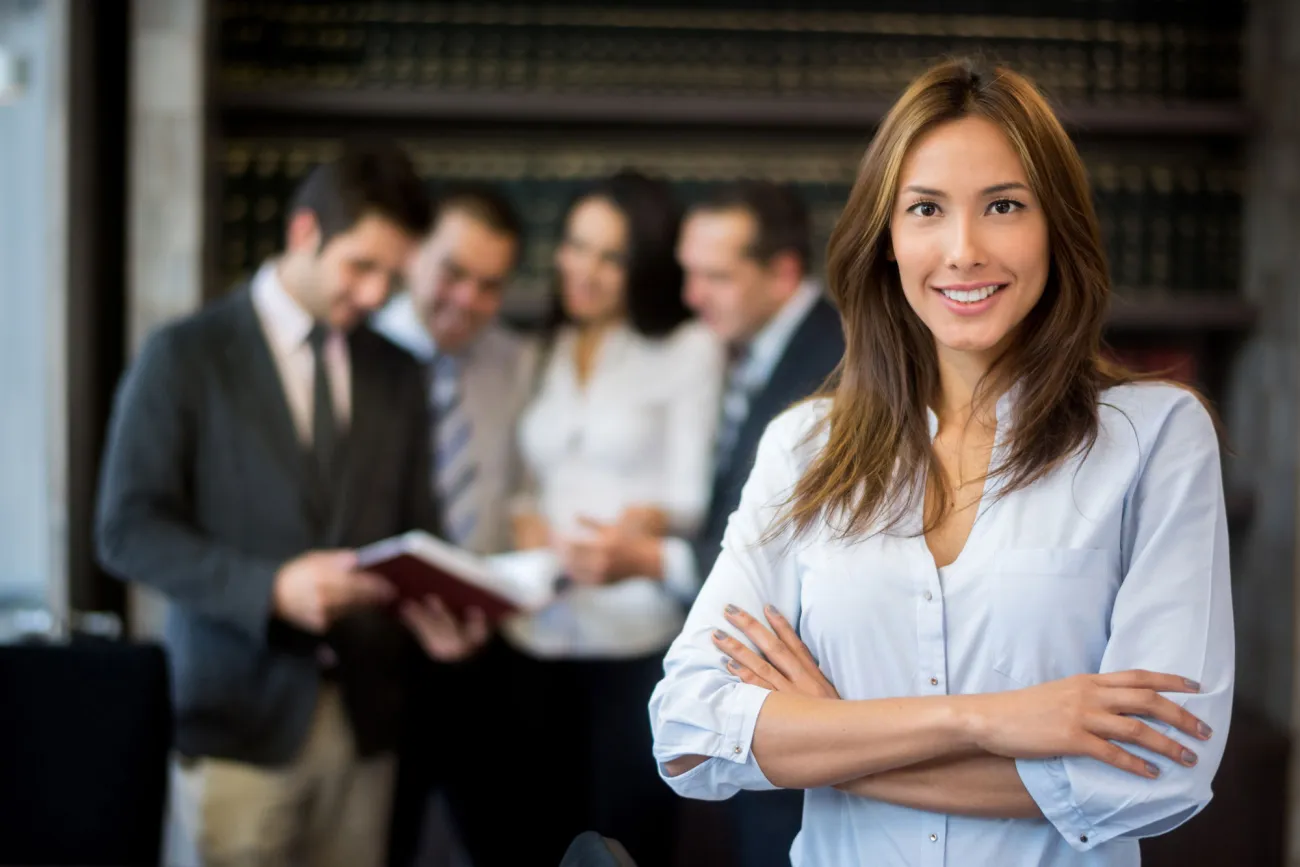 A lawyer smiling with her arms crossed with a group of lawyers examining a document in the background.